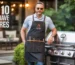 Man wearing a black grilling apron with multiple tool pockets standing beside a stainless-steel gas grill on a backyard patio.